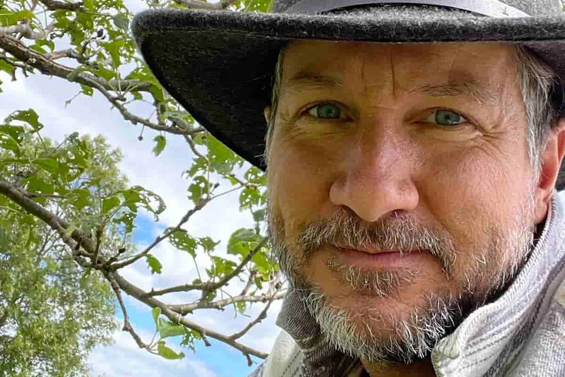 David All in a felt hat under the branches of an apple tree on his family's heritage orchard